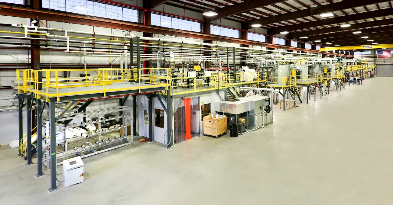 A series of machines and work areas is arranged in a line inside a large manufacturing space with an open, concrete floor and a high ceiling. Running along the array of machines are several platforms connected by walkways and stairways. 