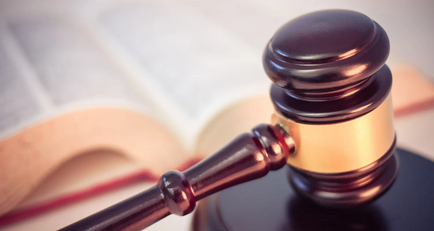 Photo of a brass and wooden gavel laying on a round wooden base with an open book in the background.