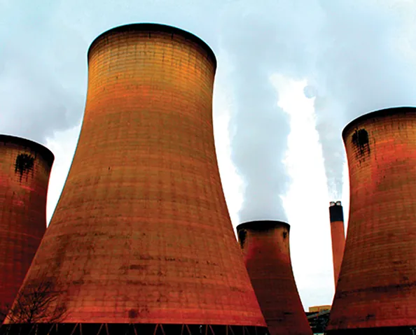 A low-angle shot captures several large, reddish-brown industrial cooling towers, with visible steam emanating from some of them and a tall, slender smokestack in the background, all set against a cloudy, light grey sky.