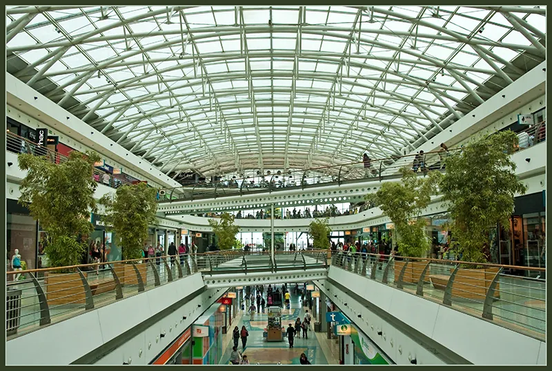 People are walking through a multi-level shopping mall. The stores are visible on the sides and the roof is made of glass.