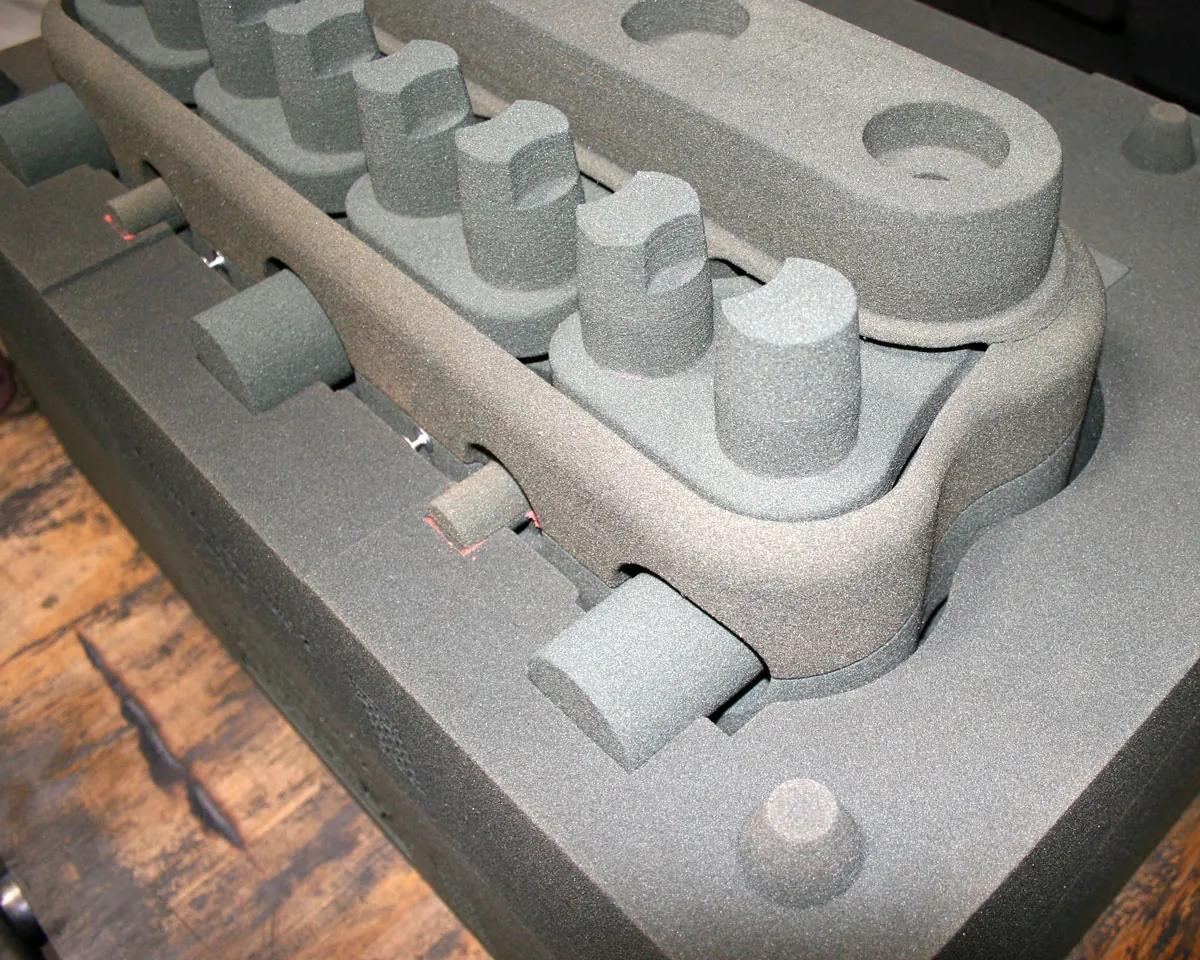 A sandy textured mold of an engine block. A number of paired columns sit inside a larger curved structure, and several cylindrical passageways emerge horizontally from the main body.