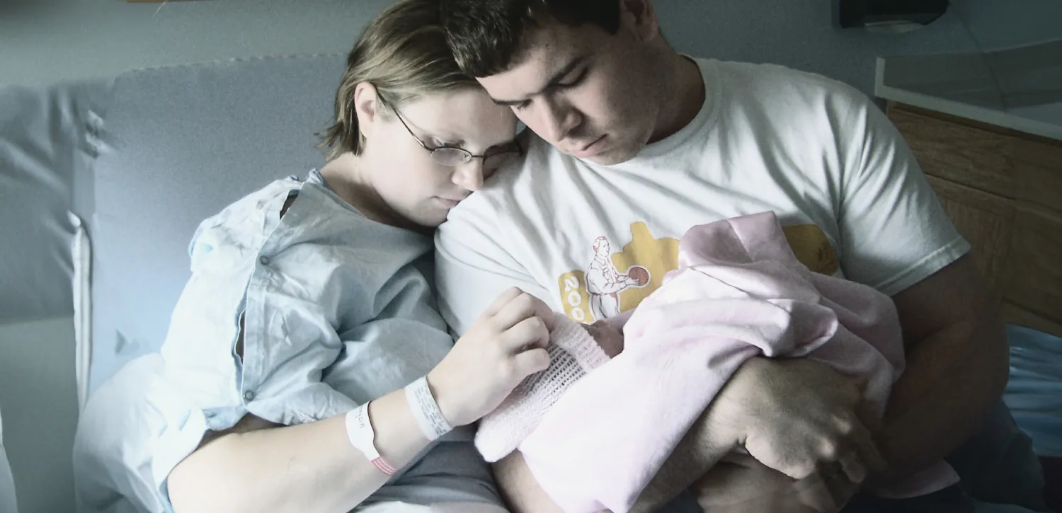 Parents hold a deceased infant in their arms in a hospital bed.