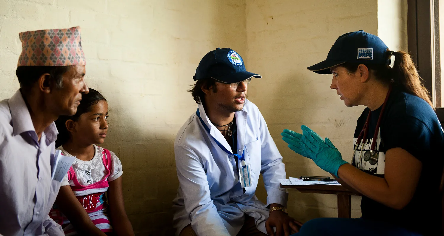 A healthcare worker wearing latex gloves sits across from three people. She speaks to the person closest to her while the other two look on.