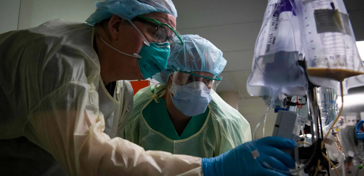 Photo of two gloved and masked medical professionals adjusting medical flow rates on a machine in a medical setting.