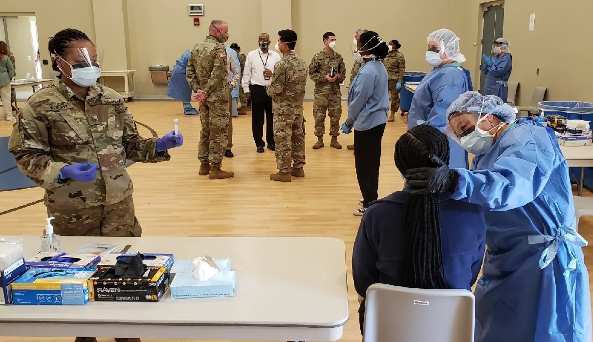 A medical worker in scrubs holds the back of a seated person's head while a uniformed National Guard member stands nearby holding testing equipment.