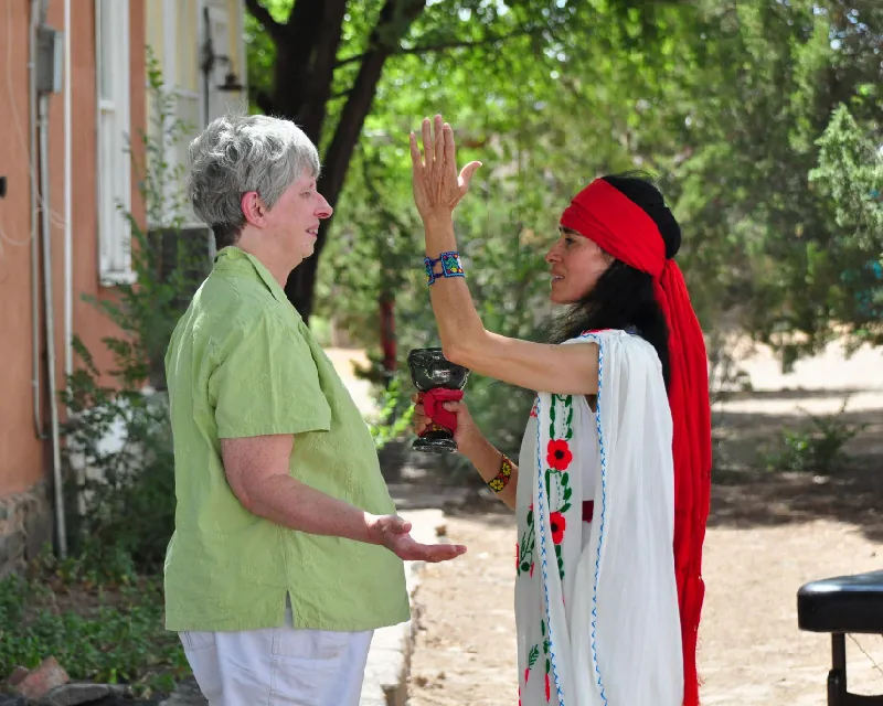 Two people stand facing each other outdoors. One stands with their hands open near their hips. The other wears a flowing garment and a long scarf tied around their forehead and stands, one hand raised in front of their head and the other holding a chalice.