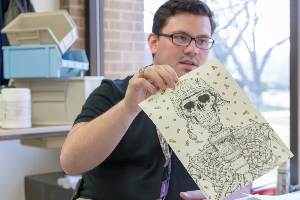 A person holds up a drawing of a skeleton wearing a combat helmet surrounded by prescription pills.