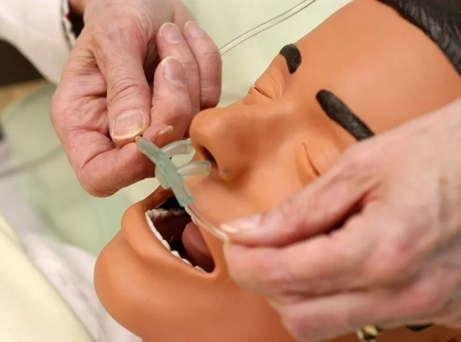 The image shows a close-up of hands placing a nasal cannula on a medical training mannequin. The mannequin has a realistic appearance, with closed eyes, defined facial features, and an open mouth. The nasal cannula, a device used to deliver supplemental oxygen, is being carefully positioned into the mannequin’s nostrils. The hands are gently holding the cannula and its tubing, demonstrating proper placement technique.