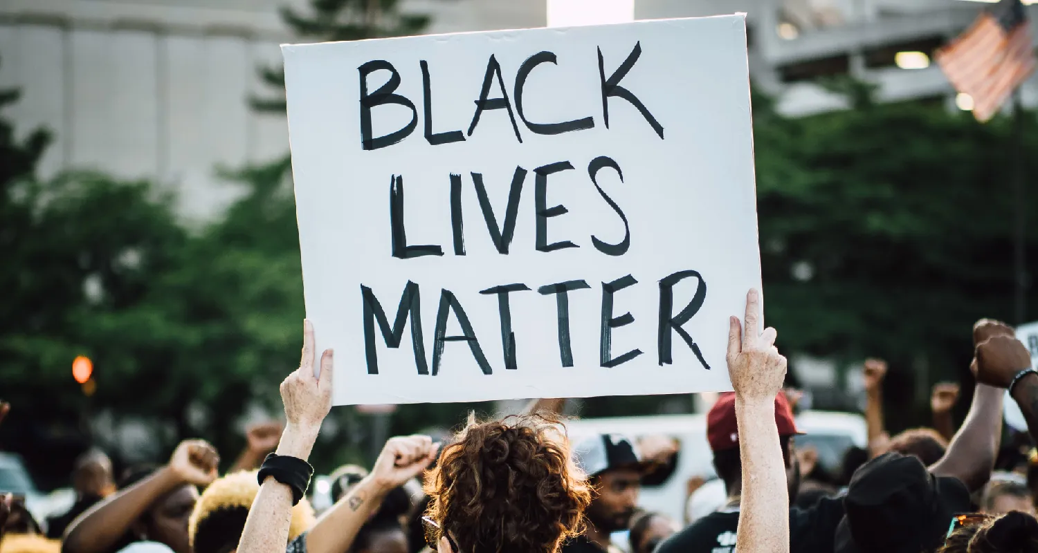 A crowd of people stand in a group outside. One person holds a sign over their head that says Black Lives Matter.
