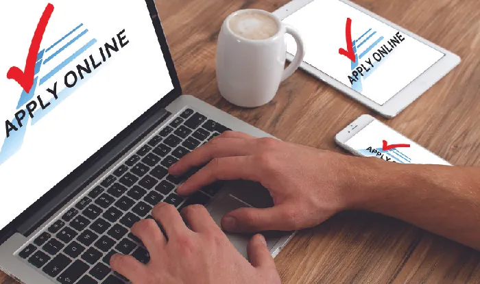 A photograph shows a computer and the hands of a person. The screen on the computer says “Apply Online.”