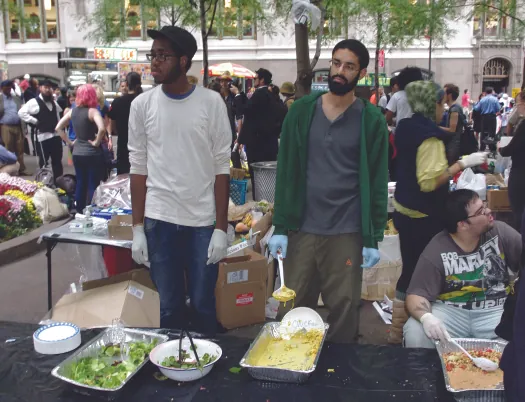 An image of three people behind a table. On the table are serval large open containers of food. A crowd of people is in the background.
