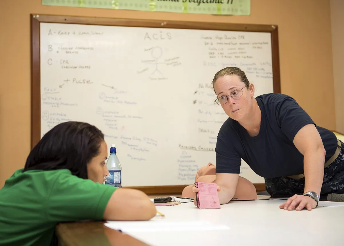 Photo of military personnel leaning over a table to get closer to student (also leaning over table) while teaching. Whiteboards in the background depict information on ACLS.