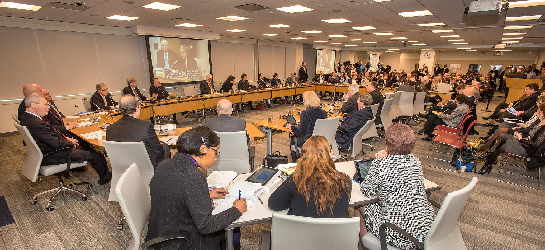 A conference hall where a large number of people are sitting and working on paper and tablet computers. A smaller group is seated around a circular table, while others are watching a video on screen in audience-style seating.