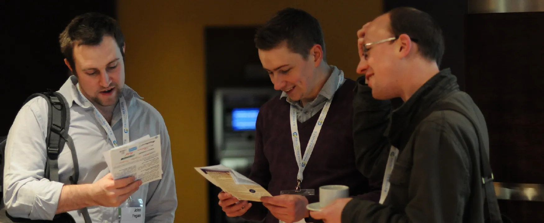 Three men at a professional event, including John Fegan, review materials and chat, all wearing 'PAIDONRESULTS' lanyards. One man smiles, holding a coffee cup, while others examine documents.