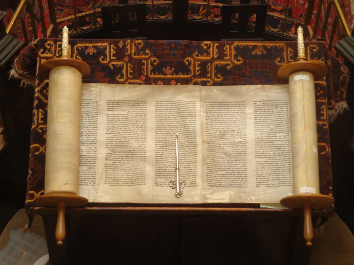 Photo of an old scroll, rolled up on both sides, opened up on an ornately decorated podium. A shiny, thin, object is laid out in the middle of the scroll.