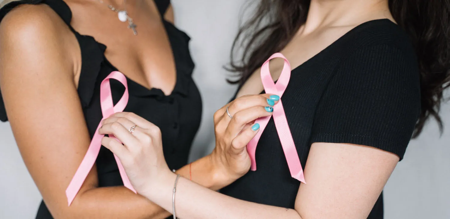 Photo of two female torsos in black dresses holding pink ribbons on each other’s breasts.
