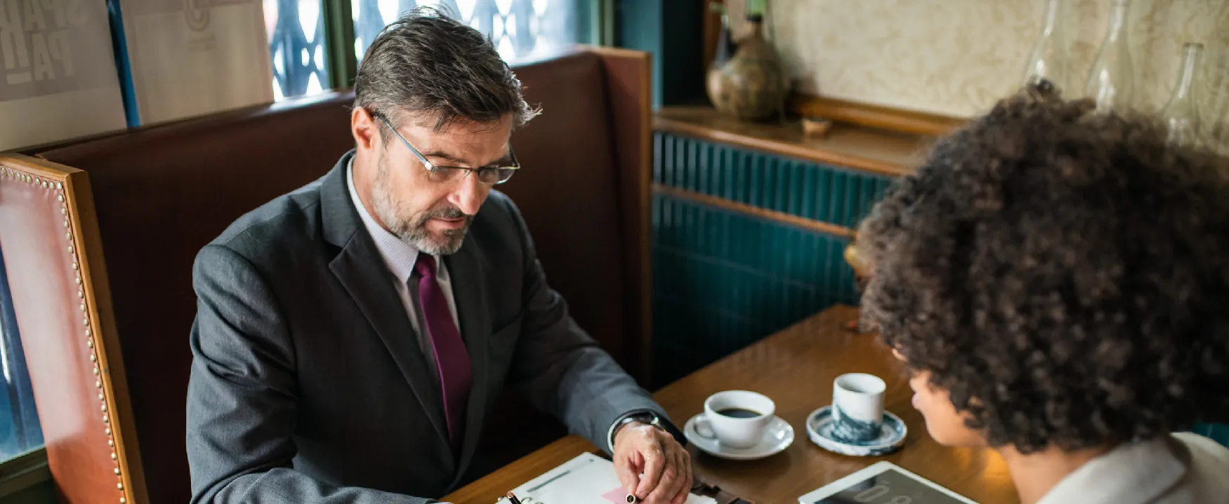 Two professionals discuss business over coffee in a cafe, with the man in a suit reviewing documents while the woman listens intently across the table.