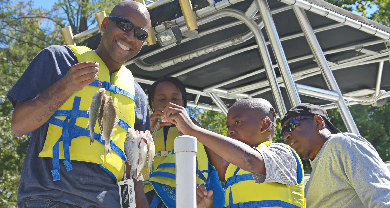 Two adults and two children stand on a small boat. Three of the people wear life vests. Two hold up a string of small fish.