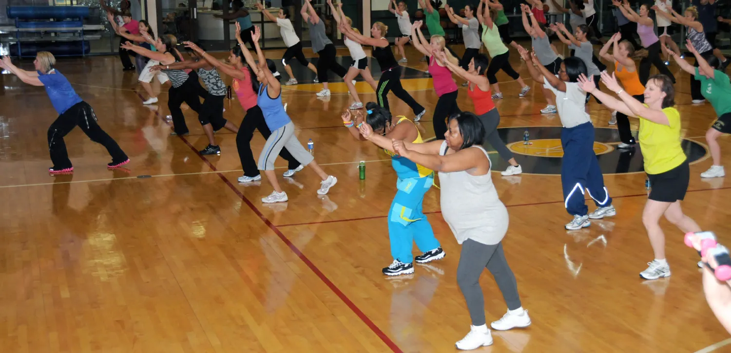 Image of people participating in a Zumba class with their arms outstretched in front of their heads, led by an instructor at the left.