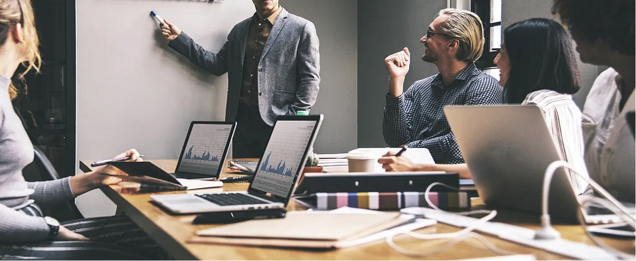 Photo of people sitting around a conference table using laptops, while someone stands at a whiteboard.