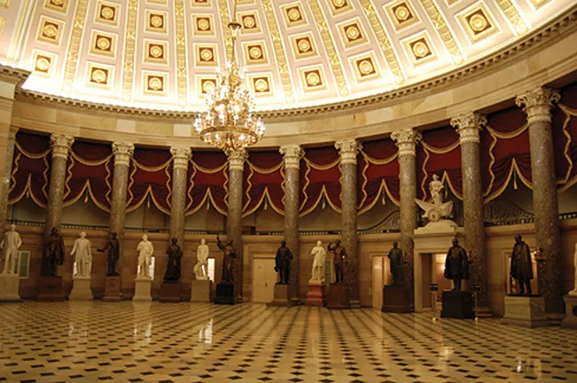 The grand National Statuary Hall in the US Capitol, featuring a magnificent dome, ornate chandelier, marble columns, red drapes, and statues of prominent figures on a checkered floor.