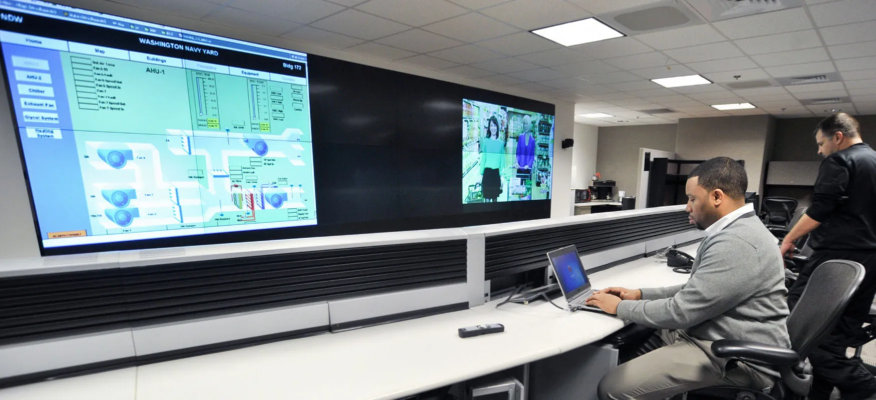 An individual works on a laptop while a large screen shows on a wall.