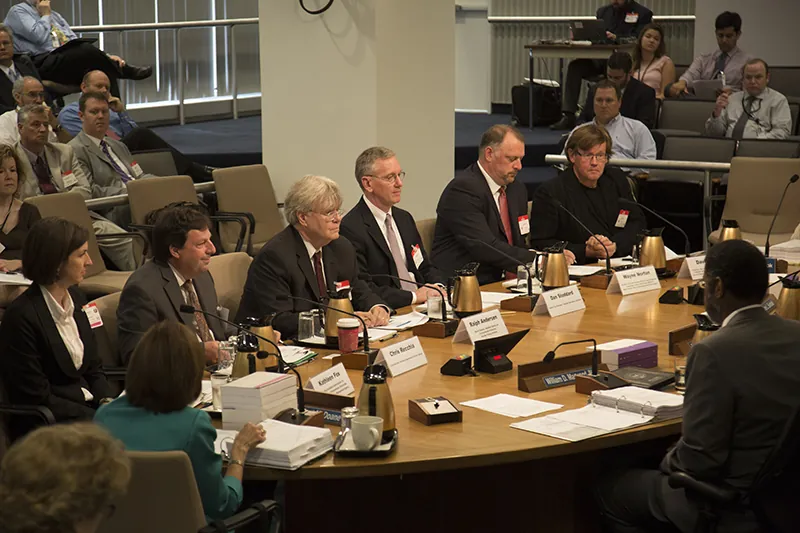 A group of officials sit around a conference table, surrounded by other seated observers who look on.