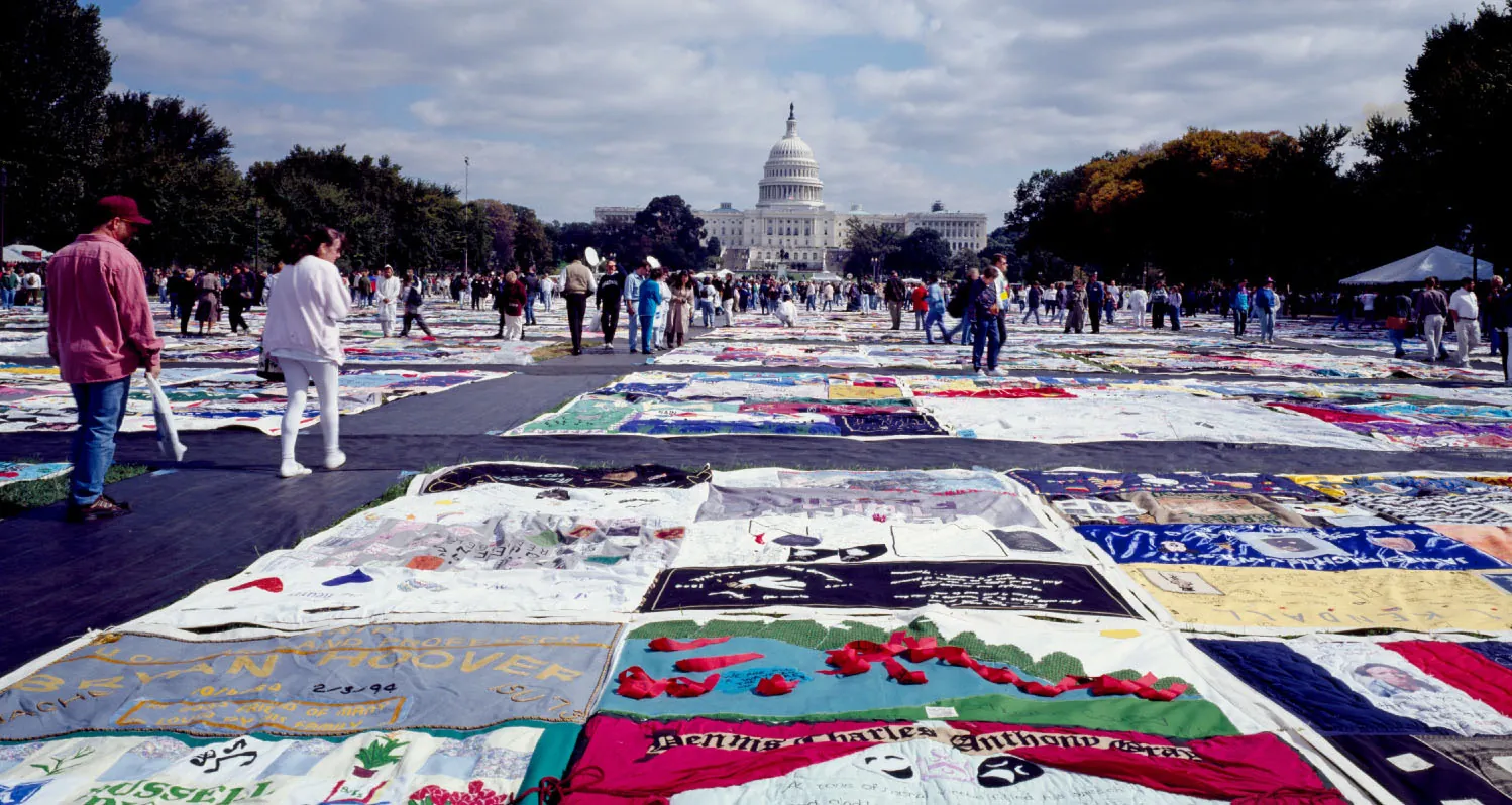 People explore a large, colorful quilt displayed at the National Mall in Washington, DC. The quilt features numerous panels, each with unique designs and messages, commemorating individuals who died of AIDS.