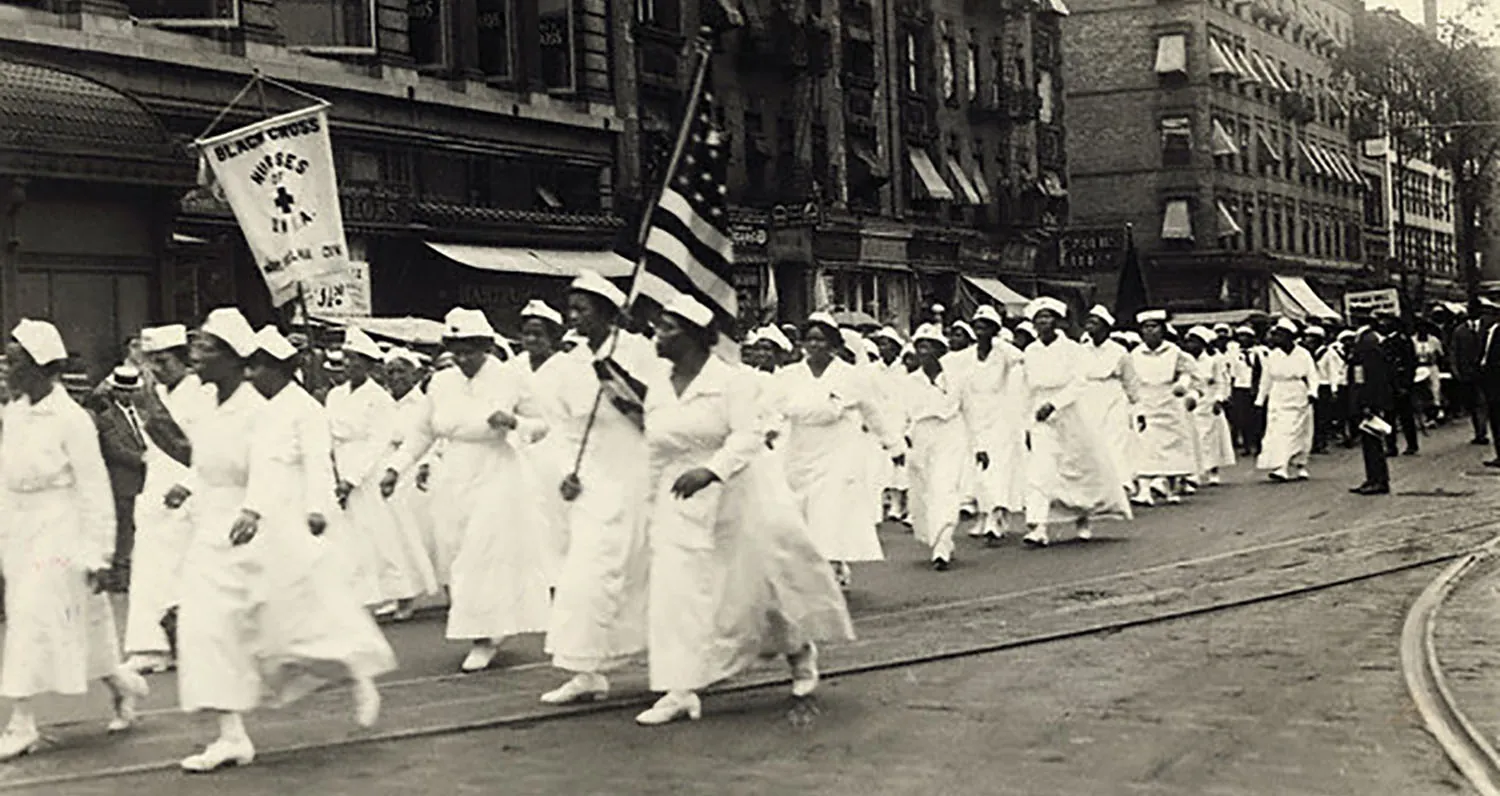 A black and white photograph of African American female nurses marching through the streets of Harlem in 1922. They are holding American flags and signs for the Black Cross.