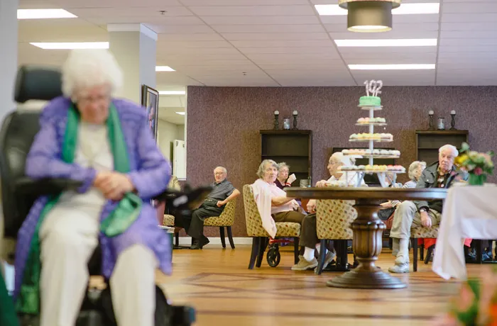 Photo of older individual sitting alone and off to the side in a room filled with other individuals sitting at tables.