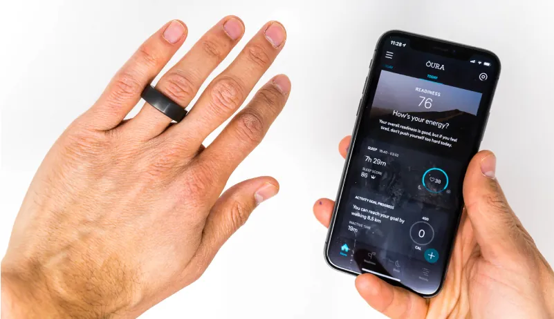 A left hand is shown with a black, large ring located on the ring finger. On the right, a hand is holding a cell phone showing health tracking information on the screen.