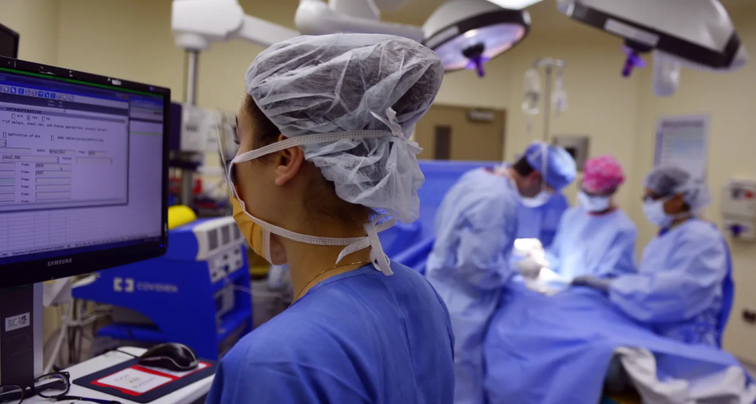 A photograph of a nurse watching a computer in the operating room, with other medical providers performing a surgery in the background.