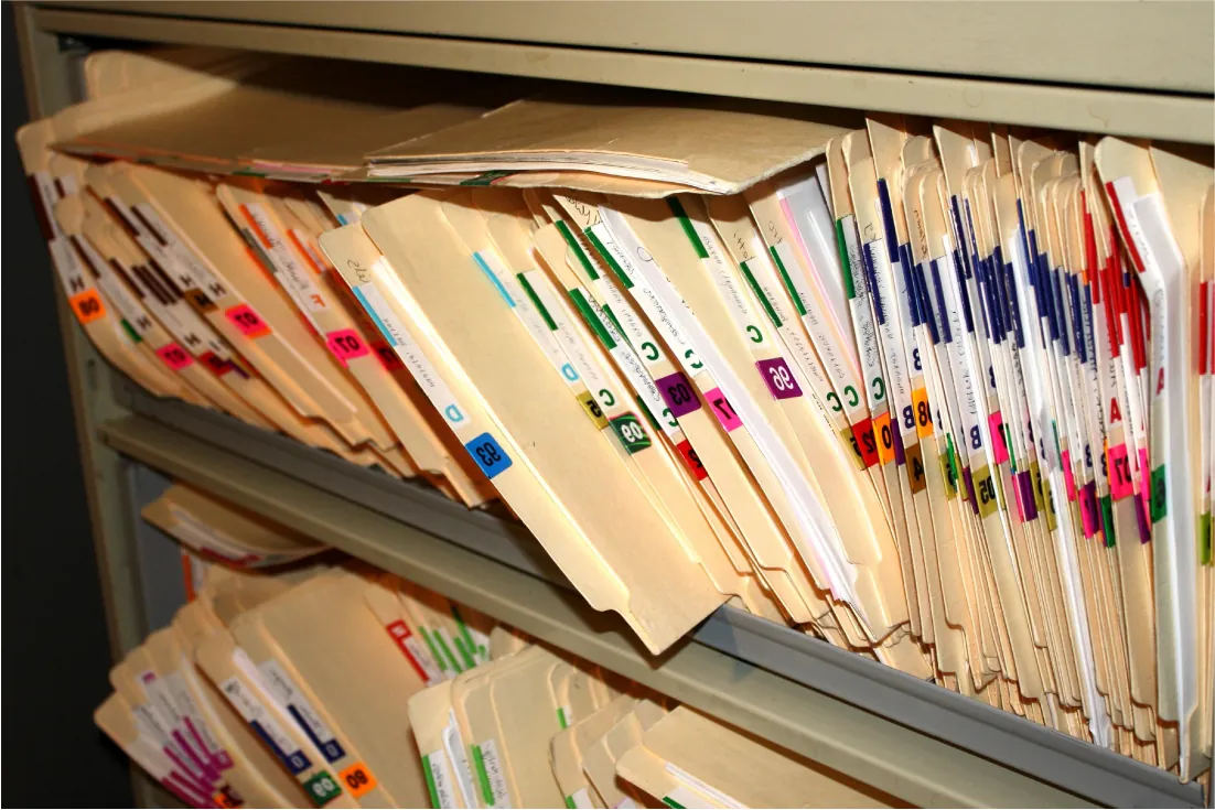 An image shows metal shelves filled with paper-filled file folders. The visible sides of the folders display tabs with various colored tabs of letters, colors, and numbers.