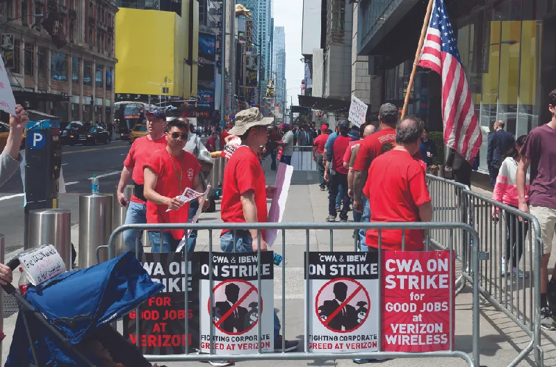 This image shows a group of people dressed in matching red shirts in an area enclosed with signs that say “CWA on strike for good jobs at Verizon Wireless” and “CWA and IBEW on strike. Fighting corporate greed at Verizon.”
