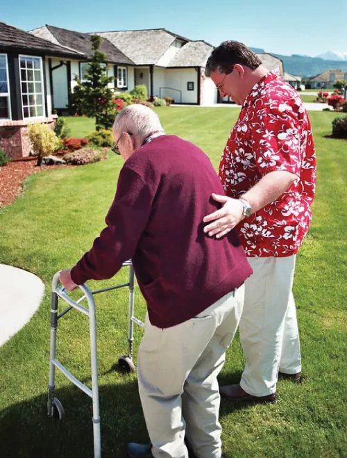 An elderly man uses a walker with the help of a caregiver.