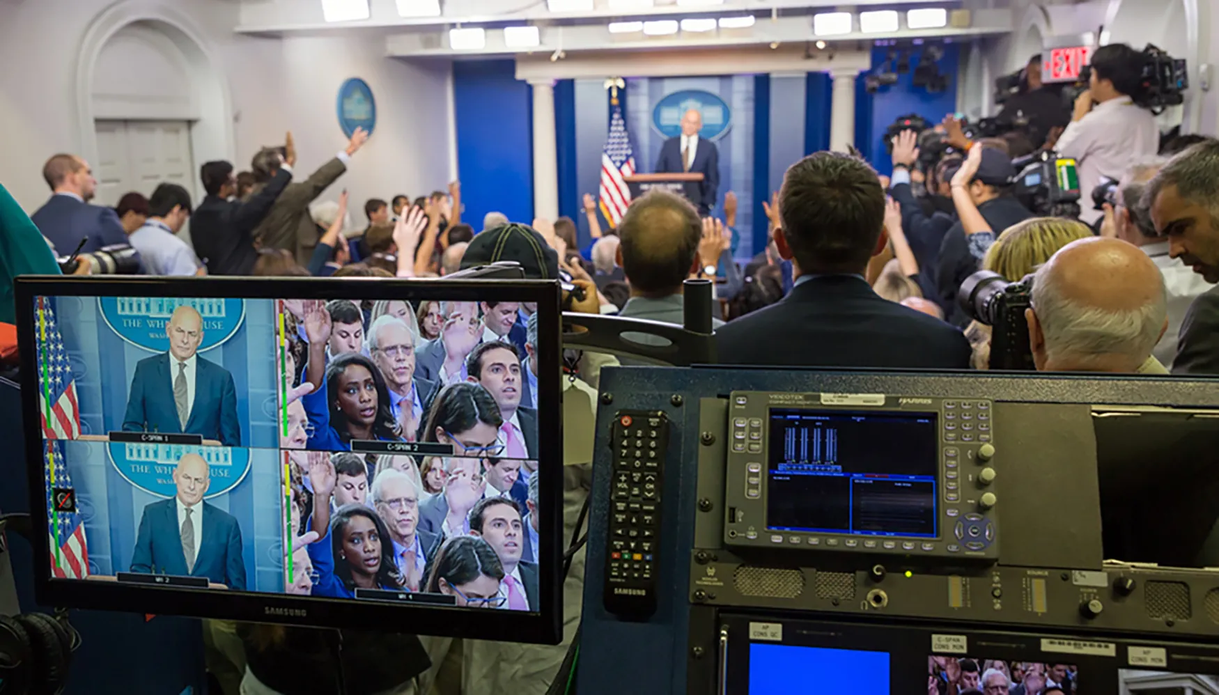 A close up view of a television monitor and control panel inside the White House briefing room. On the screen are two television feeds, and the control unit has additional smaller screens as well as switches and a keyboard. In the background the White House press corps and is visible and the Chief of Staff stands at the podium.