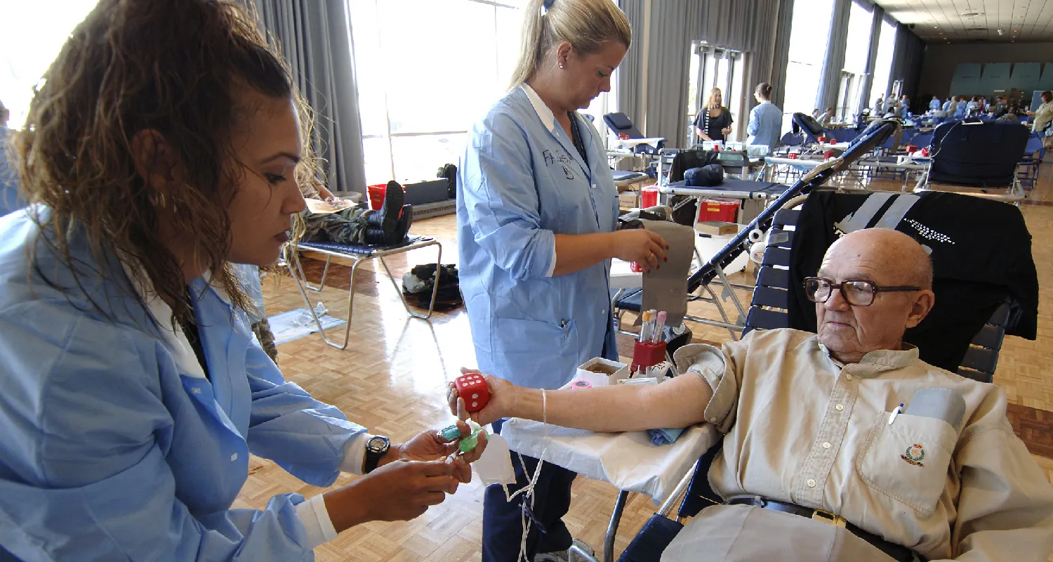 A nurse prepares to draw blood from a seated older client while another nurse stand working nearby.
