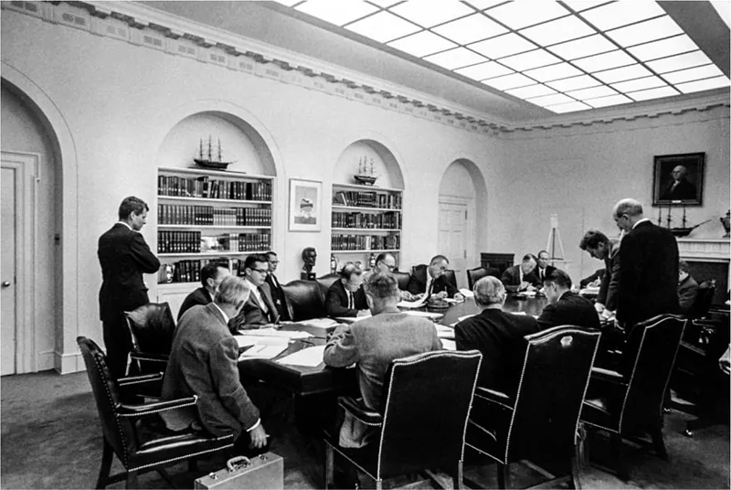 A group of officials wearing suits work with note pads and other papers around a conference table. Most are seated, although three are standing. A portrait of George Washington hangs on the wall in the background.