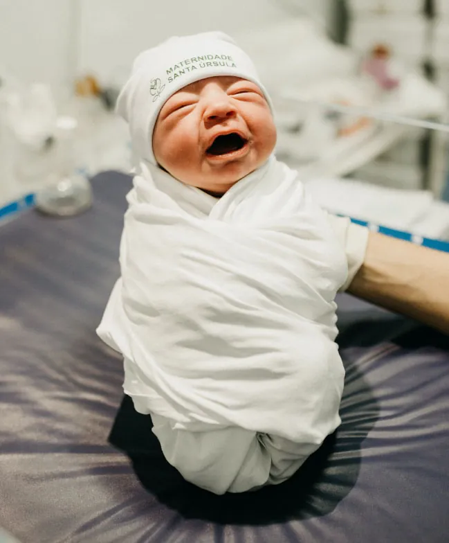 A newborn baby, swaddled in a white blanket and wearing a hat from 'Maternidade Santa Ursula,' lets out a loud cry after birth.
