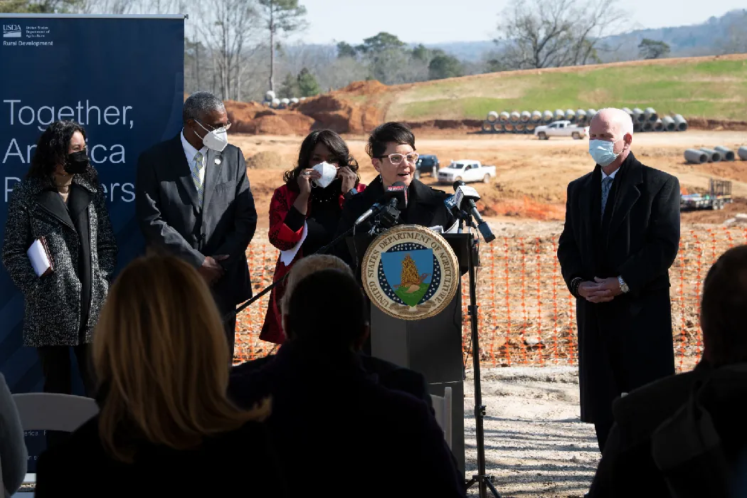 A person speaks to an audience outdoors at a podium with an insignia on its front. Several people stand listening in the background. The land behind them has been cleared and leveled for construction.