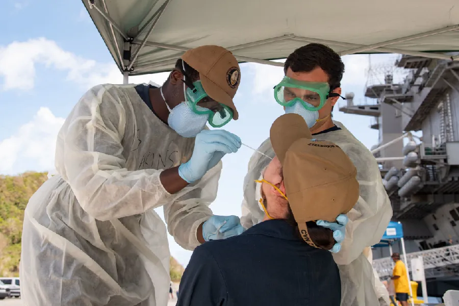 Two medical personnel stand under a tent outside, wearing protective gowns, face masks, and goggles. One swabs the nose of a person seated in front of them while the other holds the back of the seated person's head.