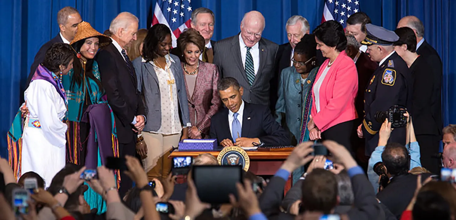 President Obama sits at a table signing a document as people stand behind him and look on. A large group of people are in front holding cameras.