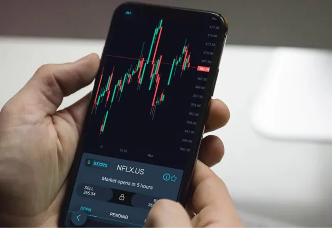 A person holds a phone. The screen reads NFLX.US, market opens in 5 hours, and shows a bar graph of stock prices.