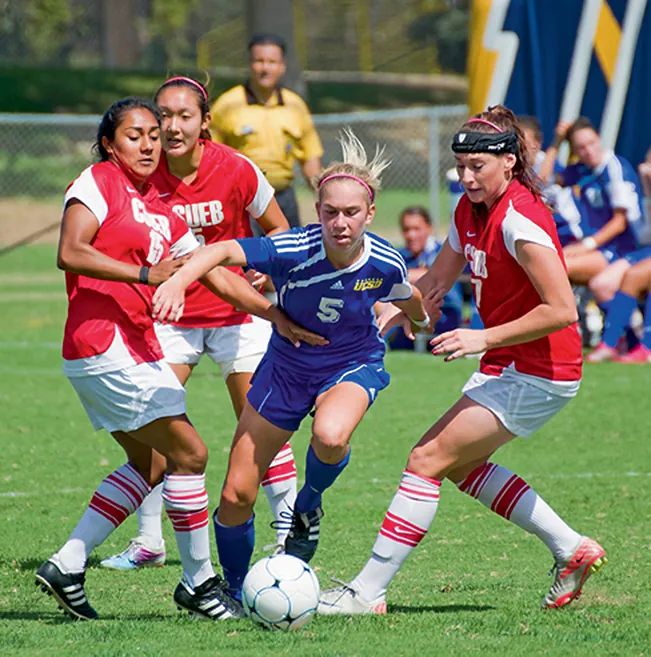 Intense soccer action unfolds as a player in a blue jersey fights for possession, surrounded by opponents in red and white. The ball is at her feet in a pivotal moment of the game.