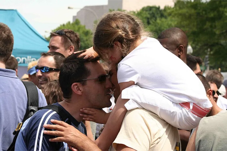 Photo of a same-sex couple kissing with child on shoulders of one of the parents.