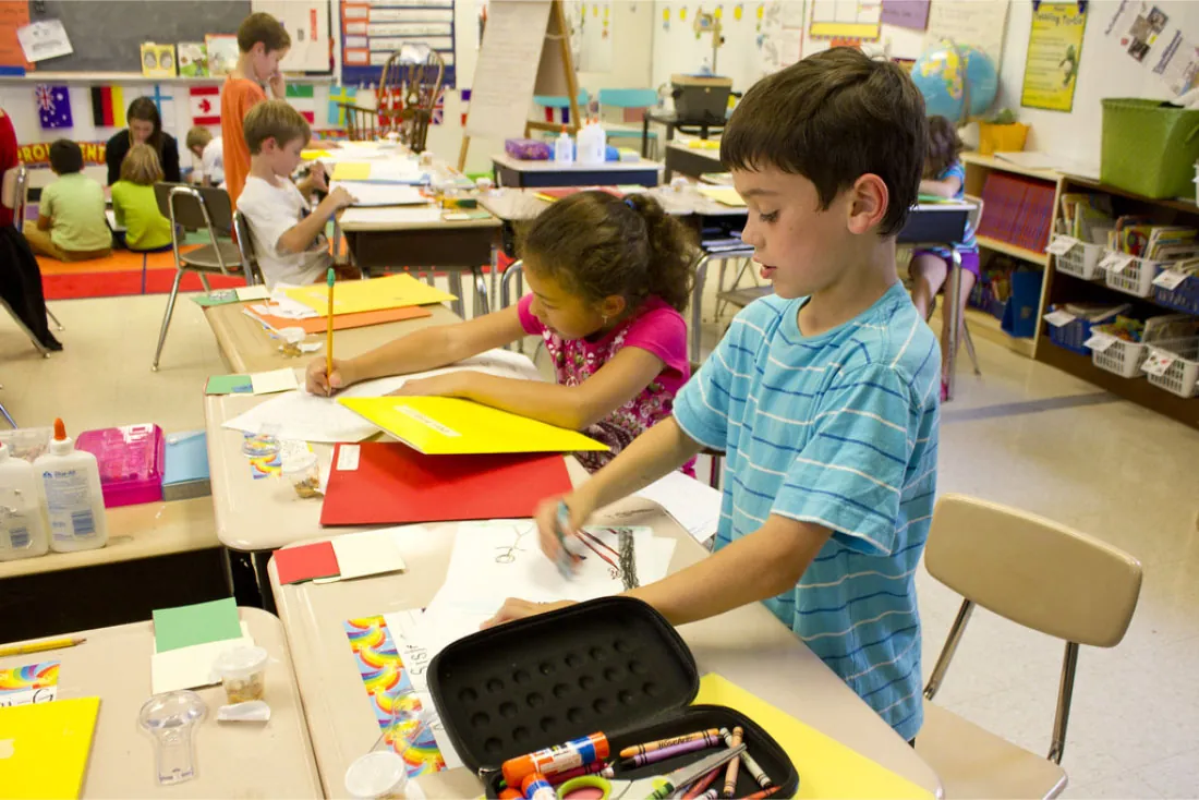 Photo of classroom of young children coloring and writing at desks. Some are sitting with a teacher on a rug.