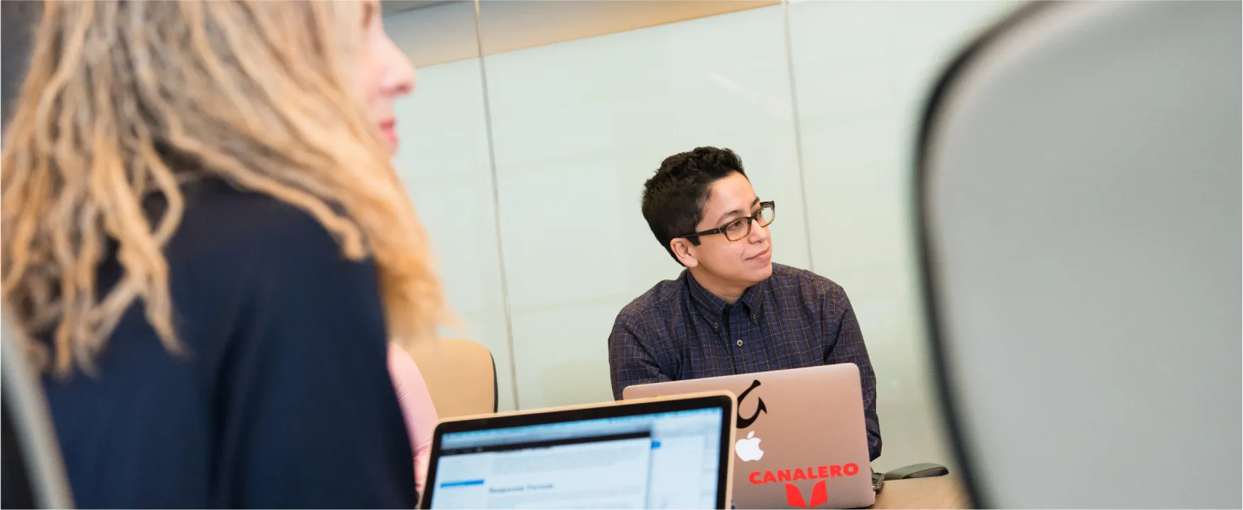 Two people are shown looking up toward the front of a large meeting table with laptops open in front of them.