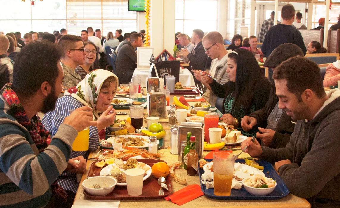 A roomful of diverse people sit at long tables, eating a meal together.