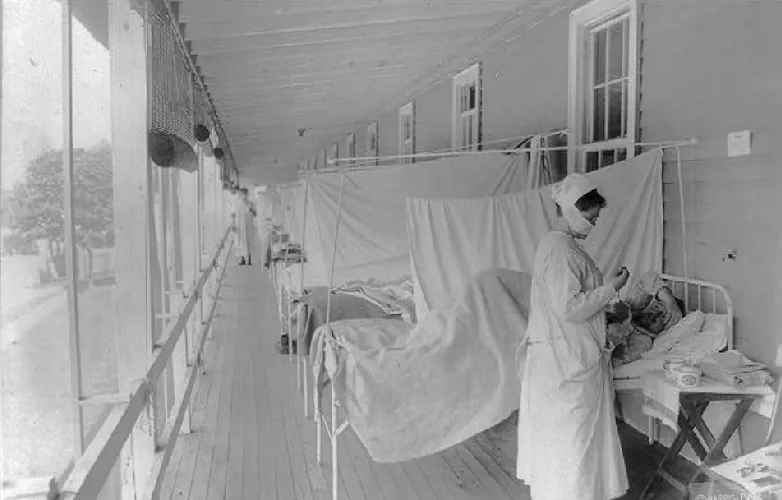 A nurse stands outside on a porch with a cloth covering over her nose and mouth. Behind her is a row of metal cots separated from one another with a hanging sheet.