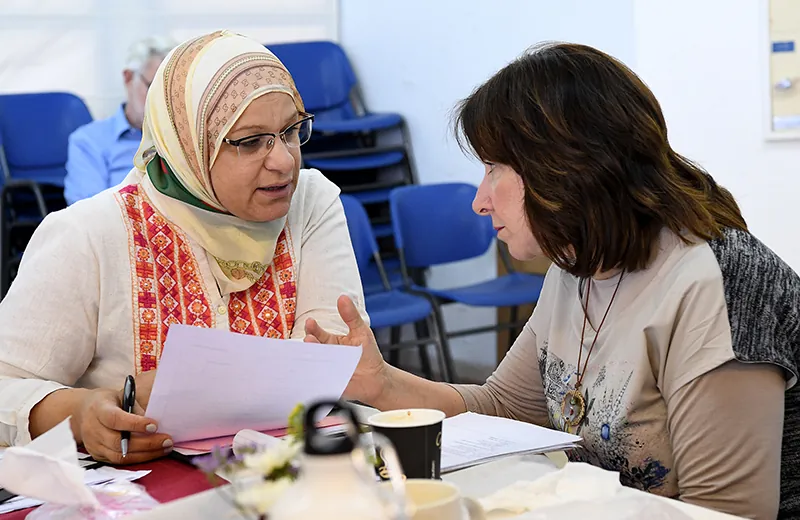 A photo shows two people sitting at a table and discussing something while holding papers. 